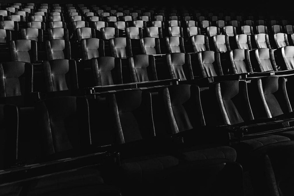fekete-fehér fotó, üres mozi. A moziterem vörös és fekete foteleit egy fénysugár, egy alacsony kulcs világítja meg. Szelektív fókusz. Karantén, korlátozó szalag a székeken  - Fotó, kép