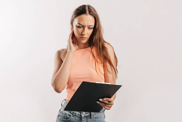 Girl student frowns with displeasure and reads information in documents. Beautiful young business woman holds a clipboard with mockup on gray background. Study and business concept. Online learning - Zdjęcie, obraz
