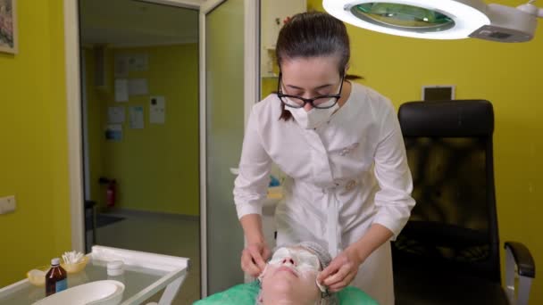 Kosmetikerin in Schutzmaske wischt Gesichtsmaske auf der Haut junger Frau mit Wattepads ab - Filmmaterial, Video