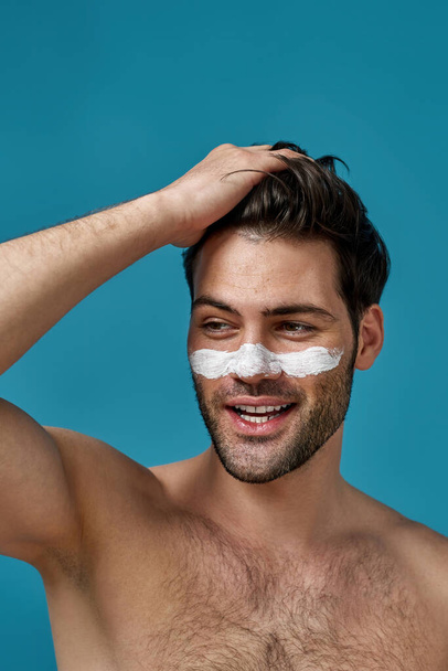 Sexy shirtless guy with white mask applied on nose for cleansing pores smiling aside, touching his brown hair isolated over blue background - Fotografie, Obrázek