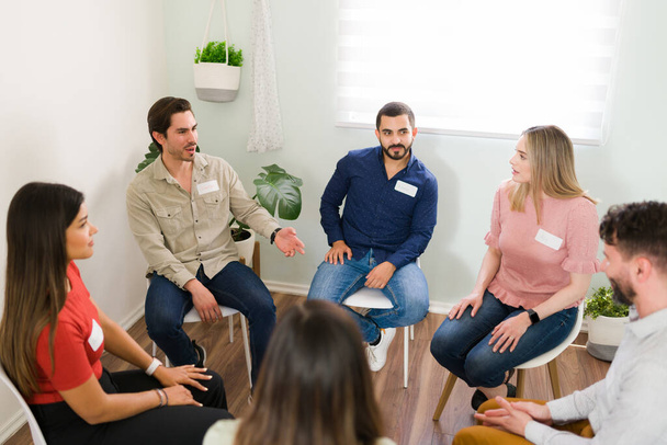 Młodzi ludzie rozmawiają podczas sesji terapii grupowej. Różnorodna grupa kobiet i mężczyzn dzielących się swoimi problemami i proszących o poradę w zakresie zdrowia psychicznego - Zdjęcie, obraz