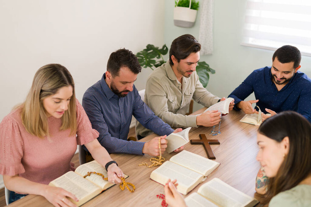 Разнообразная группа целеустремленных молодых людей, читающих свои молитвы с католическими четками во время изучения Библии вместе  - Фото, изображение