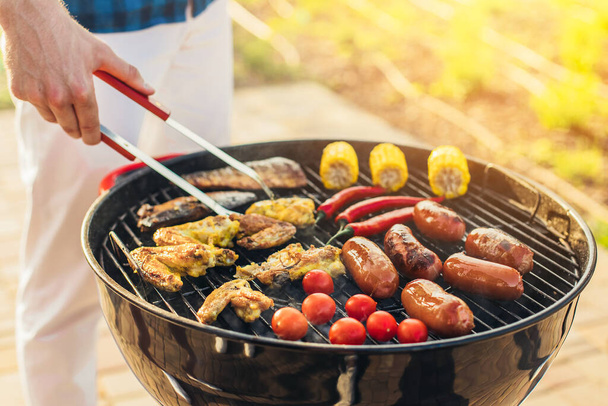 Man making barbecue, assorted vegetables and chicken wings with sausages, grilling on a portable barbecue outdoors in a park in nature - Photo, Image