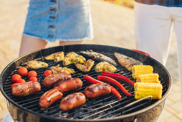 Assortment of fresh healthy vegetables, meat and sausages grilled on a hot fire on a charcoal grill, in a green grassy spring or summer field with copy space - Photo, image