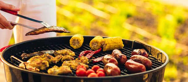 Man fries meat and vegetables on the grill, flips sausages with tongs, corn cobs, chili peppers and chicken wings outdoors in summer - Fotoğraf, Görsel