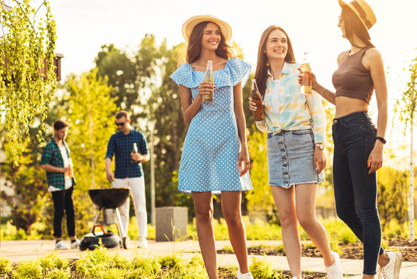 Three beautiful girlfriends having fun on a picnic, drinking drinks and chatting, a group of young friends doing barbecue on the grill in the park at sunset - Фото, изображение