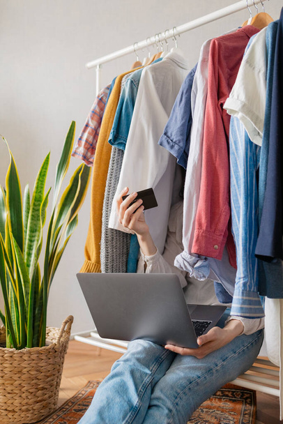 white European woman sits in Wardrobe, among the clothes and makes a purchase online, woman with a laptop and phone orders a new dress and other clothes in the online store - 写真・画像