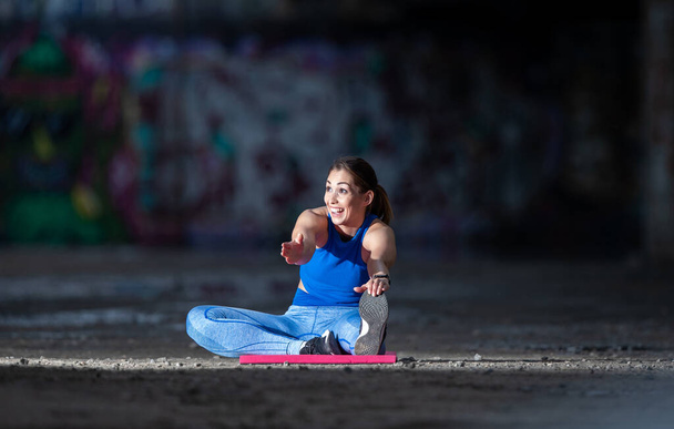 座って一本足の曲げウォーミングアップストレッチを行うスポーティーな女の子。落書きと壁の前の都市部で運動する若い選手.  - 写真・画像