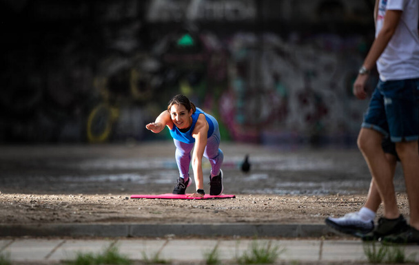 Jeune femme sportive faisant de l'exercice à l'extérieur en zone urbaine avec des gens qui passent par là. Fit girl faisant planche à un bras dehors au soleil.  - Photo, image