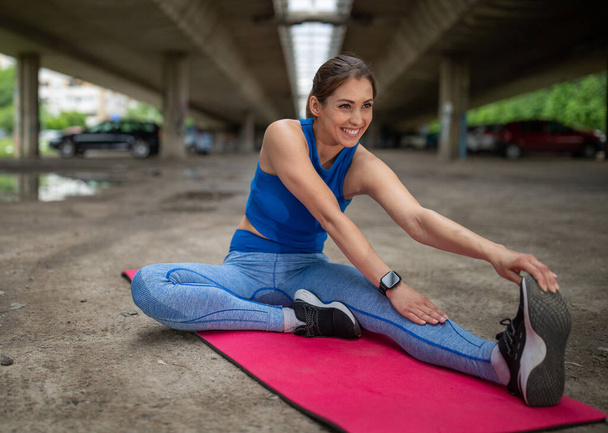 Flexibele jonge vrouw doet zittende eenbenige bocht naar voren. Atleet sporten glimlachen buiten op roze mat buiten. - Foto, afbeelding