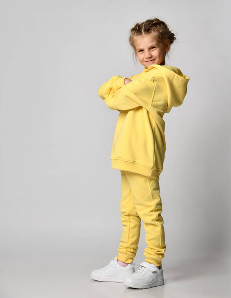 active little girl folded her arms over her chest and looks at you. place for text. Happy cute child in a yellow tracksuit on a white background. - Фото, изображение