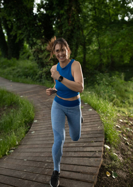 Fit girl running outdoors in park on path. Young athletic woman smiling exercising wearing sportswear and smartwatch. - Photo, Image