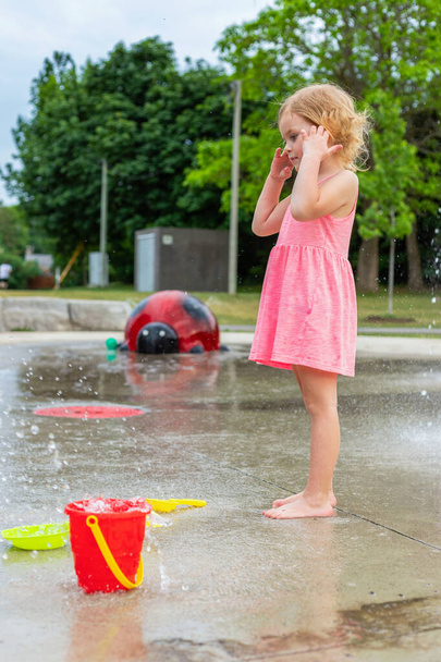 Μικρό παιδί παίζει με το νερό και τα παιχνίδια στο splash pad στο τοπικό δημόσιο πάρκο κατά τη διάρκεια της ζεστής ημέρας του καλοκαιριού. Μικρό όμορφο κορίτσι σε ροζ φόρεμα διασκεδάζει στην παιδική χαρά σιντριβάνι. - Φωτογραφία, εικόνα