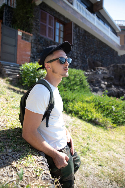 A young attractive caucasian man is wearing sunglasses and backpack, it is summer and the day is beautiful, hipster with hands in pockets, traveler concept. - Фото, зображення