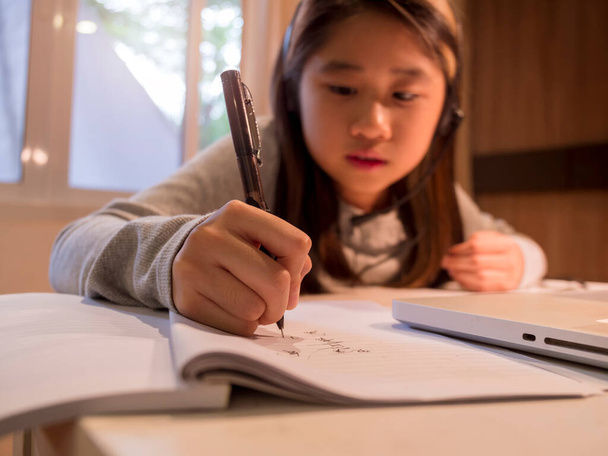 Kulaklıklı Asyalı kızın eli bilgisayarın başında masa başında çalışıyor. Çocuk, internetten karantina dersleri alarak not defterine kulaklık takıyor. Kız öğrenci covid pandem nedeniyle okuldan sanal İnternet öğreniyor - Fotoğraf, Görsel