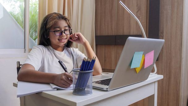  Asian girl in headphones sit at desk study online on laptop. Kid wear headset handwriting in notebook learning using internet lessons on quarantine. Girl student learning virtual internet online class from school due to covid pandemic. - Photo, Image