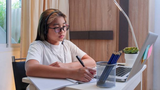  Азійська дівчина в навушниках сидить за комп'ютером на ноутбуку. Малюк носить навушники з почерком у ноутбуці, використовуючи уроки інтернету на карантині. Дівчата, які навчаються віртуальному інтернету онлайн-класу зі школи через ковалентну пандемію.. - Фото, зображення
