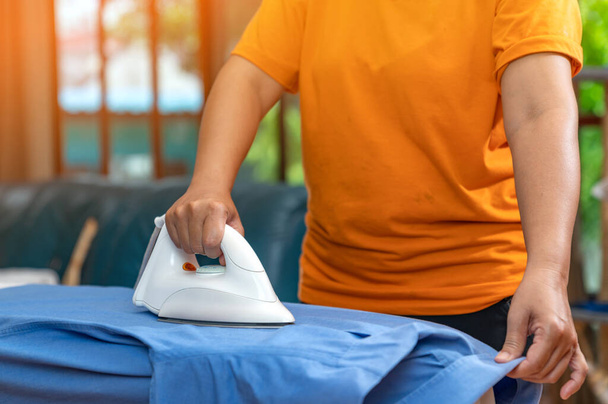 Camisa naranja de utensilios de mujer planchando camisa azul sobre tabla de planchar en la sala de estar. - Foto, imagen
