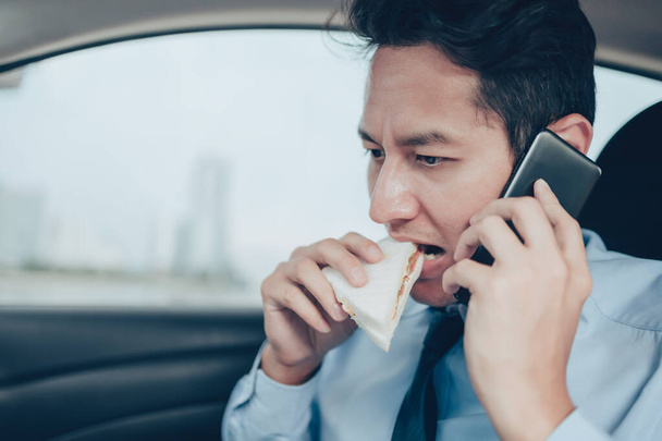 Geschäftsmann isst Sandwich, während er auf dem Fahrersitz seines Autos telefoniert. Geschäftiger Geschäftsmann und Essen im Auto - Foto, Bild
