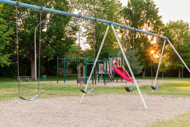 Aire de jeux sans enfants dans le parc public local, balançoires sur les chaînes avec du sable en dessous, herbe, arbres, structure de jeu et glissades rouges en arrière-plan dans la soirée d'été avec coucher de soleil. - Photo, image