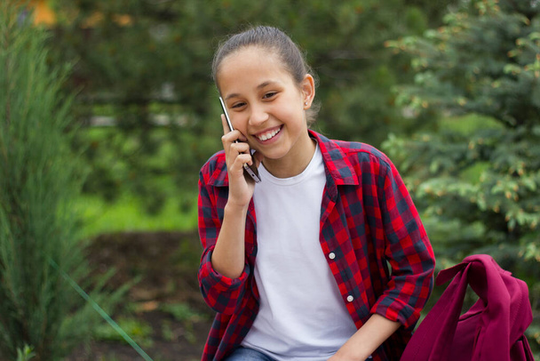A sad girl with long dark hair in a white T-shirt and a plaid shirt with a backpack sits in the park enthusiastically uses the phone to communicate in the messenger. Selective focus - Photo, Image