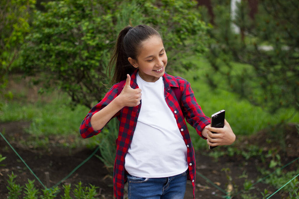 A cheerful girl with long dark hair in a white T-shirt and a plaid shirt in the park enthusiastically uses the phone to communicate in the messenger take a selfie and show cool class. Selective focus - Photo, Image