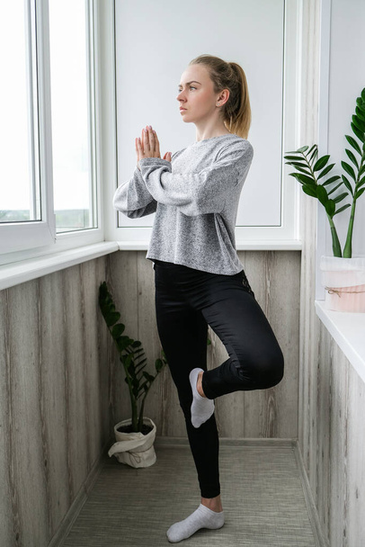 Young millennial blonde woman doing yoga exercise stretching fitness at balcony home. Mindfulness meditation. Relax breathe easy pose gym healthy lifestyle concept. - Zdjęcie, obraz