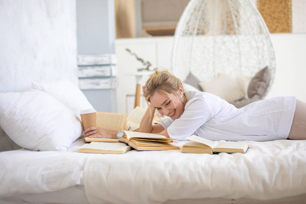 Красивая чувственная молодая женщина с светлыми волосами в белой рубашке читает книги и пьет кофе на кровати в современном доме - Фото, изображение