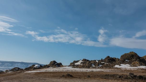 Zimowy krajobraz. W dolinie jest śnieg i sucha trawa. Na tle błękitnego nieba znajduje się pasmo górskie pozbawione roślinności. Malownicze chmury. Syberia - Zdjęcie, obraz