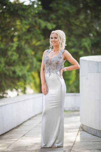 An attractive Caucasian female wearing an elegant long silver dress posing in a park - Photo, Image