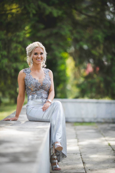 An attractive blonde Caucasian female wearing an elegant long silver dress sitting in a park - Photo, image