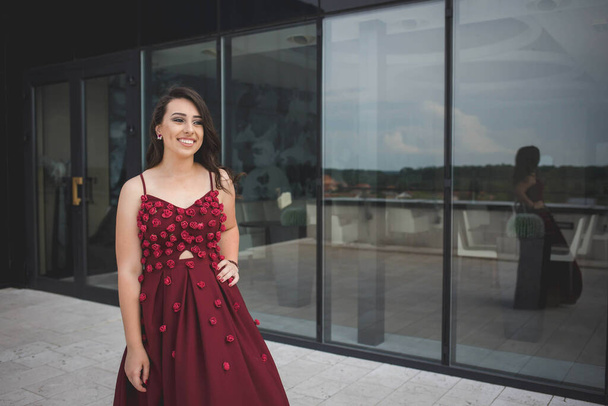 An attractive Caucasian female wearing an elegant long burgundy dress on background of a building - Photo, Image