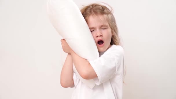 Söpö pieni takkuinen unelias tyttö halailee tyynyä valkoisella taustalla. lasten varhaiset nousut kouluun ja lastentarhaan. mukava vuodevaatteet. - Materiaali, video