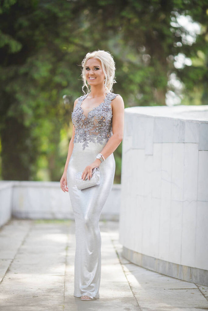 An attractive Caucasian female wearing an elegant long silver dress posing in a park - Photo, image