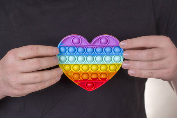 Um jovem tem um coração de arco-íris nas mãos. Nova moda colorido Silicone antistress Pop It Toy. - Foto, Imagem