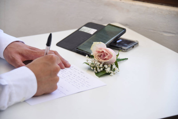 A groom writing a letter on a white paper with his boutonniere and smartphone on the table - Фото, изображение