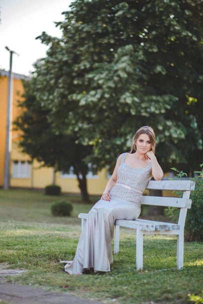 A pretty Caucasian woman with a silver evening dress and heavy makeup sitting on a wooden bench - Zdjęcie, obraz
