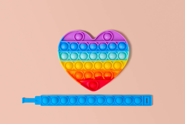 Nuevo juguete de silicona colorido de moda-antiestrés Pop It en forma de corazón de arco iris y una pulsera de silicona azul sobre un fondo beige - Foto, imagen