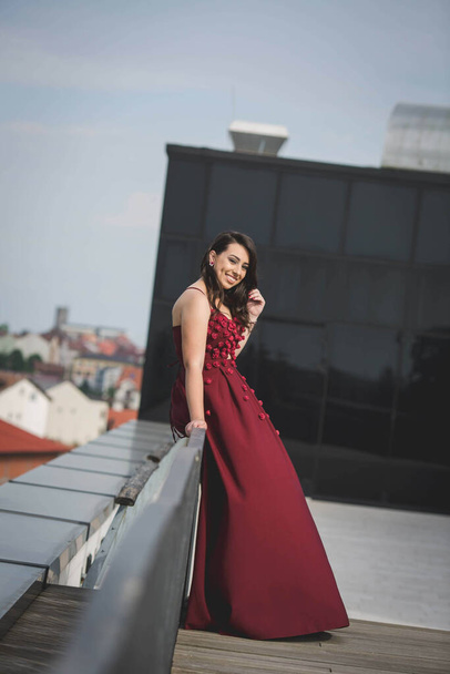 An attractive Caucasian female wearing an elegant long burgundy dress sitting outdoors - Fotografie, Obrázek