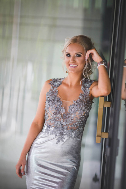 A pretty blonde Caucasian woman with a silver evening dress leaning on the door of a building - Foto, afbeelding