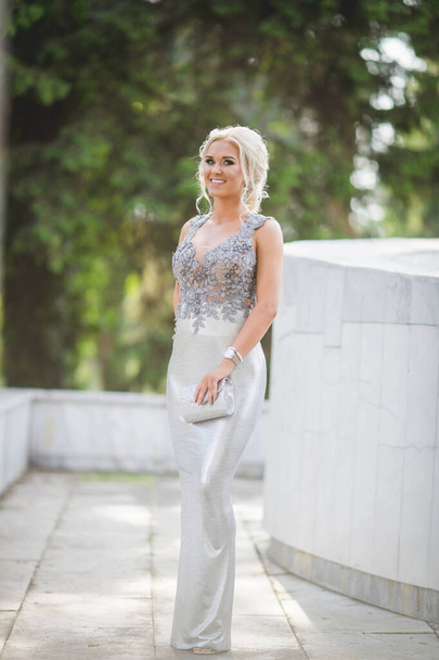 An attractive blonde Caucasian female wearing an elegant long silver dress posing in a park - Photo, Image