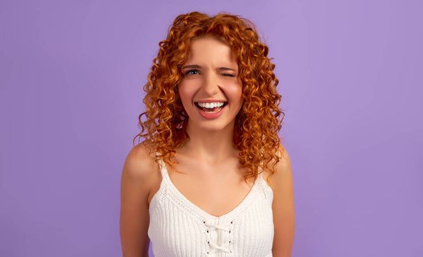 Cute redhead girl with curls winks isolated on purple background. - 写真・画像