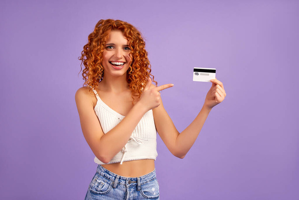 A cute red-haired girl with curls holds a credit card and points a finger at it, isolated on a purple background. - Photo, Image