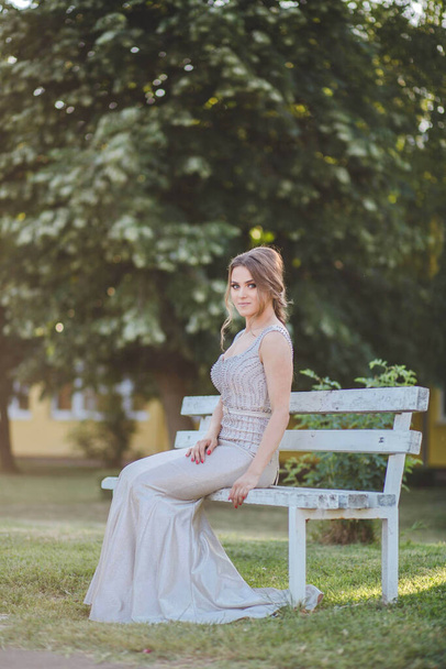 An attractive Caucasian female wearing an elegant long silver dress sitting on a bench in a park - Valokuva, kuva