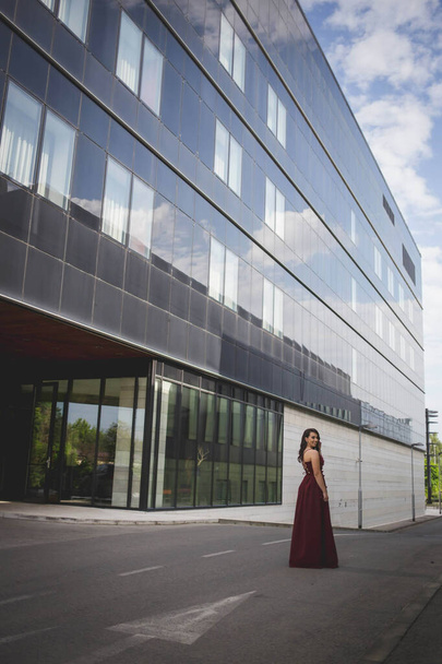 A pretty Caucasian woman with a burgundy evening dress posing in front of a hotel building - Foto, Imagen