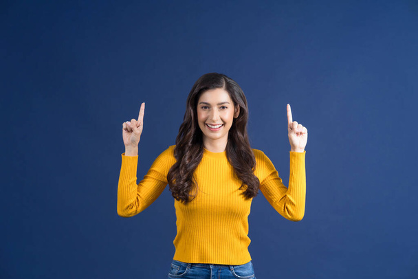 Jeune femme asiatique souriante heureuse doigt pointant vers la publicité avec une émotion heureuse sur fond de couleur bleue, montrant ou présentant le produit via le concept d'espace de copie vide - Photo, image
