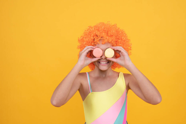 happy child in swimsuit wearing orange curly wig hair holding french macaron, sweet tooth. - Photo, Image