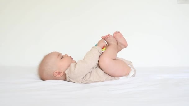Lindo bebé se encuentra en una cama blanca en la habitación y juega con un sonajero en sus manos  - Imágenes, Vídeo