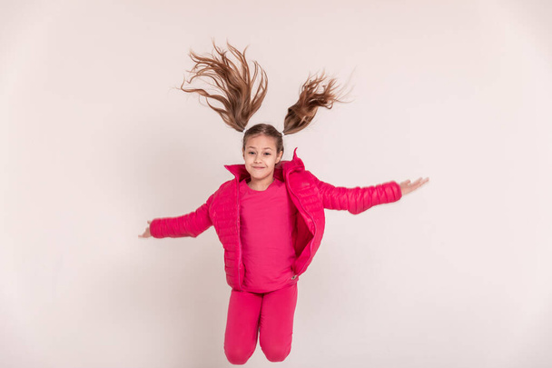 a beautiful girl in a pink jacket jumps and smiles on a white background. happy childhood. Copy space - Fotografie, Obrázek