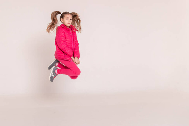 Ένα όμορφο κορίτσι με ροζ μπουφάν πηδάει και χαμογελάει σε λευκό φόντο. Χαρούμενη παιδική ηλικία. Αντιγραφή χώρου - Φωτογραφία, εικόνα